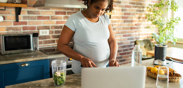 Schwangere schaut beim Kochen in den Laptop