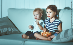 Zahlreiche Studien stellen einen erhöhten Nahrungsmittelkonsum fest, nachdem Kinder Werbung gesehen haben. Zwei Kleinkinder sitzen mit einer Schüssel Popcorn auf dem Sofa.
